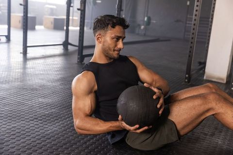 Muscular man performing russian twist holding medicine ball on rubber gym floor near squat rack