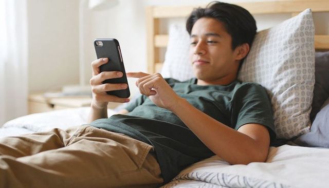 Teen relaxing on bed scrolling smartphone and tapping screen in cozy bedroom daylight