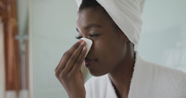 Woman in bathroom applying skincare product with a cotton pad while wrapped in white towel and robe. Great for advertisements and articles on beauty routines, wellness, self-care, and skincare products. Perfect for promoting health and beauty brands or wellness blogs.