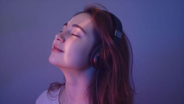 Young woman enjoying music with over-ear headphones under purple-blue gel lighting, serene