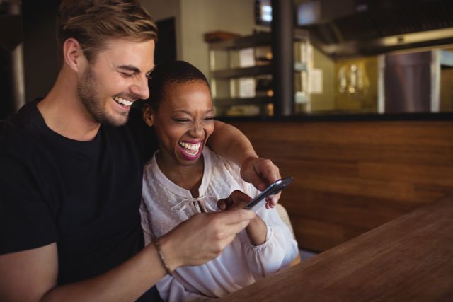 A happy couple is sitting in a restaurant, smiling and laughing while looking at a mobile phone. This image can be used to depict themes of togetherness, joy, technology use, and social interactions in a casual dining setting. It is ideal for advertisements, social media posts, and articles related to relationships, technology, and lifestyle.