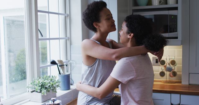 Biracial lesbian couple hugging and drinking coffee in kitchen. self isolation quality family time at home together during coronavirus covid 19 pandemic.
