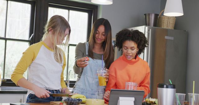 Happy diverse teenager girls using tablet, making healthy cocktail in kitchen. Spending quality time, lifestyle, friendship and adolescence concept.