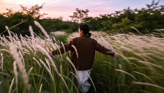 Asian woman walking through tall reed grass at sunset touching grasses with arms wide