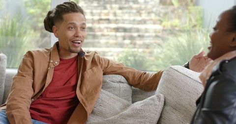 Multiracial couple chatting on light-gray sofa in bright living room with garden view