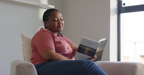 Mid-adult african american woman reading paperback in sunlit minimalist living room