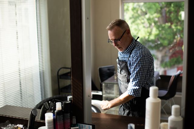 Side view of a middle aged Caucasian male hairdresser working in a hair salon, cleaning his workplace, reflecting in a mirror