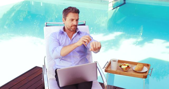 Professional man dressed in a blue shirt working on laptop by a pool with breakfast tray. Scenic outdoor setting combining business and leisure, perfect for articles on remote work, lifestyle blogs, vacation productivity or advertisements for tech and luxury services.