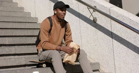 Young African American man sitting on skateboard eating lunch on urban steps in sunlight