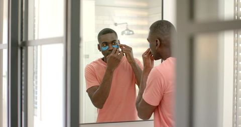 African american man applying blue under-eye patches at bright bathroom mirror