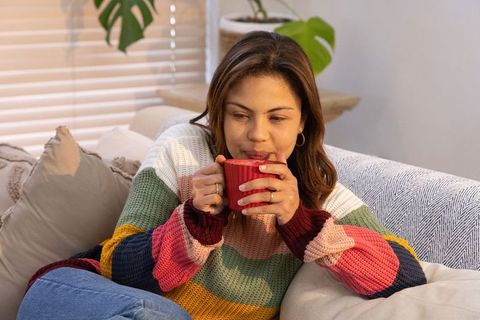 Woman sipping warm drink on sofa wearing colorful striped knit sweater, cozy living room