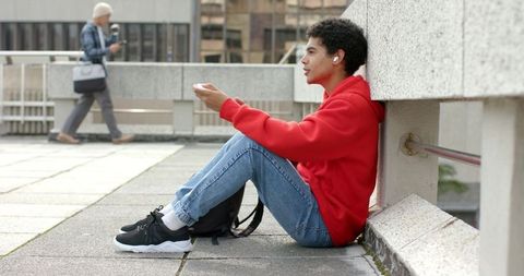 Asian young man sitting on urban rooftop in red hoodie listening with wireless earbuds