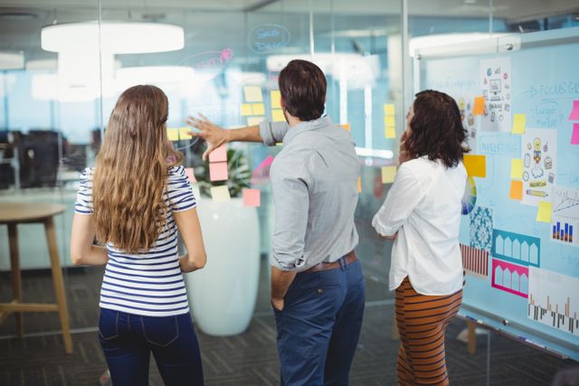 Business team collaborating on a project using sticky notes on a glass wall in a modern office. Ideal for illustrating teamwork, project planning, brainstorming sessions, and creative processes in a corporate environment.