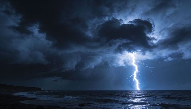 Forked lightning striking ocean horizon at night, storm clouds illuminating coastline