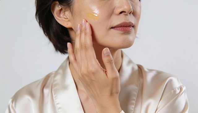 Applying beige-tinted facial serum on cheek by adult asian woman wearing satin robe
