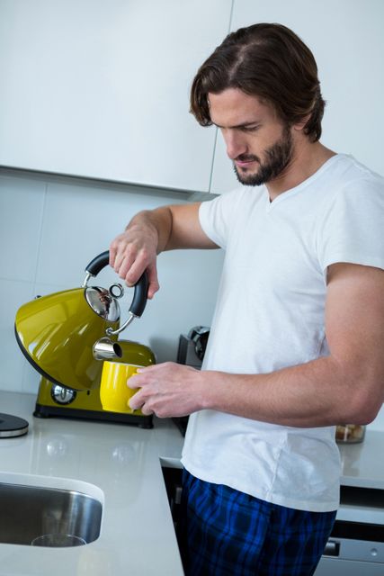Man pouring coffee into a yellow cup in a modern kitchen at home. Ideal for use in lifestyle blogs, articles about morning routines, home living, and coffee-related content. Can also be used in advertisements for kitchen appliances, coffee products, and casual homewear.