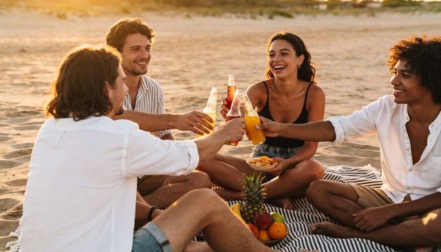 Friends toasting bottles at sunset beach picnic on striped blanket with pineapple