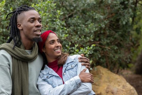 Smiling diverse couple embracing on woodland trail wearing denim jacket, scarf, headband
