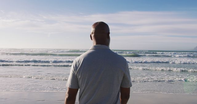 Man standing on sandy beach facing ocean waves under clear blue sky. Ideal for themes of solitude, relaxation, or mindfulness. Perfect for travel brochures, advertisements about wellness retreats, and websites promoting beach holidays.