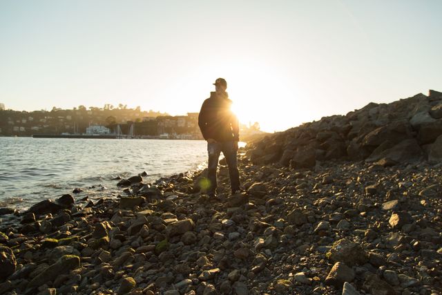 Man standing on rocky shore during sunset with sunflare in background, creating tranquil and serene mood. Ideal for use in campaigns focusing on travel, adventure, nature, relaxation, and mindfulness. The scenery can also be used for backgrounds or wallpapers for various creative projects.
