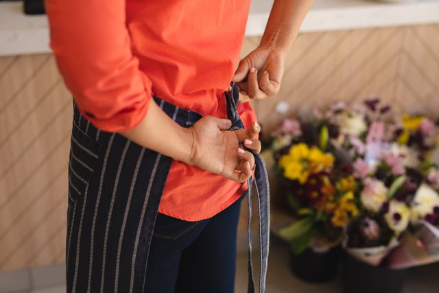 This image depicts a female cafe owner tying her apron in a flower shop. Ideal for use in articles or advertisements related to small businesses, entrepreneurship, cafes, and flower shops. It can also be used in content focusing on women in business, preparation for work, and the daily activities of a barista.
