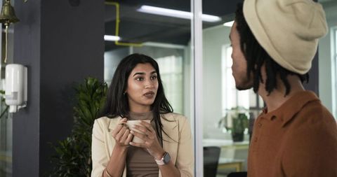 Diverse coworkers chatting in modern glass-walled office during coffee break