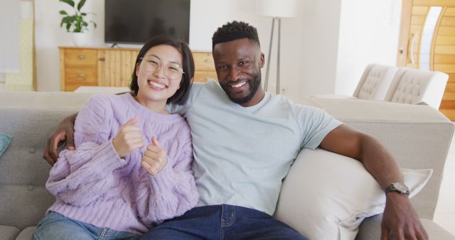 Interracial couple enjoying time together sitting on couch in modern living room. Ideal for promoting concepts of happiness, love, diversity, modern living, and relationship bonding in lifestyle blogs, advertising, or community-focused initiatives.