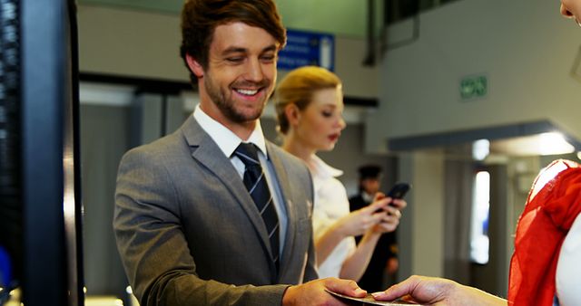 Businessman in a suit receiving boarding pass from airline staff at an airport counter. Woman holding phone in background suggests busy travel environment. Ideal for illustrating business travel, airport services, and customer service interactions.