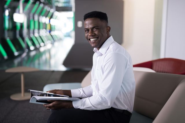 Male executive in a modern, futuristic office environment, smiling while reading a magazine. Ideal for use in business and corporate materials, advertisements for office spaces, technology and innovation-related content, and professional lifestyle promotions.