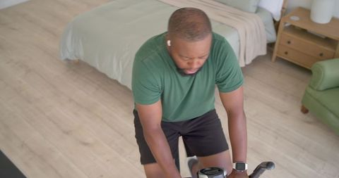 African american man exercising on spin bike in bedroom wearing smartwatch and earbuds