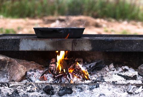 Cast iron skillet cooking over open campfire on metal grill with glowing embers and smoke