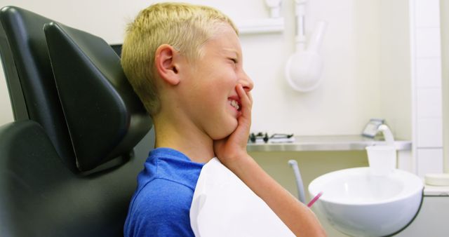 Young blonde boy sitting in a dental chair holding his jaw in pain at a clinic. He wears a blue t-shirt and appears unhappy. Suitable for healthcare, dental, or childhood images. Useful for illustrating dentist visits, dental pain, and oral health issues.