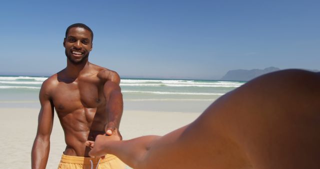 A young African American man is smiling while holding hands with someone on a sunny beach, with copy space. His cheerful expression and the beautiful coastal setting suggest a moment of relaxation and connection.