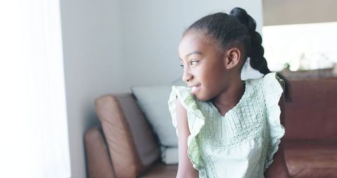 African girl gazing out window on sunlit sofa in minimalist living room, calm child portrait