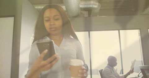 Young professional woman checking smartphone while holding coffee in modern open office
