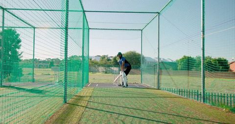 Right-handed batsman practicing in outdoor cricket net cage on sunny training pitch with protective