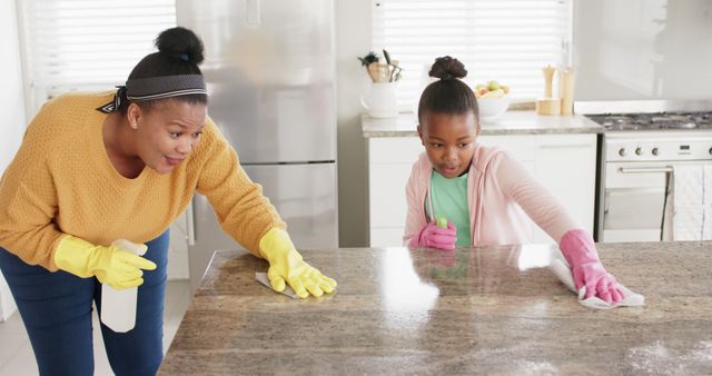 Happy biracial mother with daughter wearing rubber gloves and cleaning table in kitchen. Domestic life, family and lifestyle, unaltered.