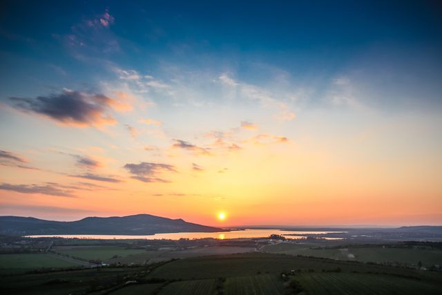 Beautiful sunset scene over a green valley with distant mountains and a tranquil lake in the background. Suitable for nature-themed backdrops, website banners, or relaxation campaign visuals. Ideal for illustrating calm and peaceful moments in the countryside.