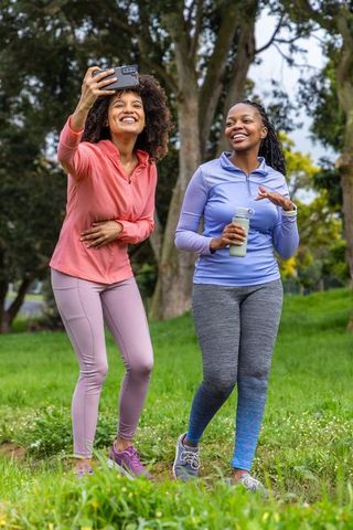 Diverse women walking in park wearing colorful activewear, taking selfie and hydrating