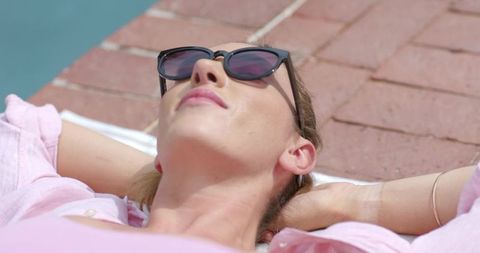 Woman sunbathing by pool wearing sunglasses and pink shirt, relaxed summer lifestyle
