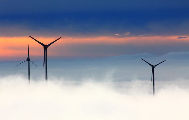 Wind turbines generating renewable energy emerge through a layer of mist at sunset, highlighting the beauty and importance of sustainable power. Ideal for illustrating concepts related to green energy, environmental conservation, and sustainable technology. Can be used for websites, presentations, and articles focusing on climate change and renewable energy solutions.
