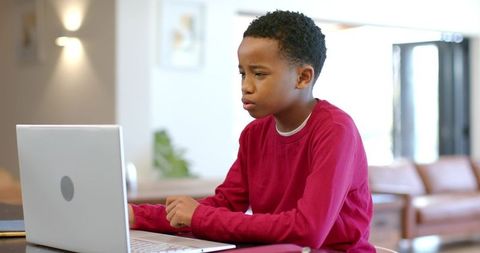 African American boy concentrating on laptop for remote learning in bright living room