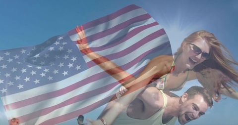 Young couple enjoying sunlit beach piggyback with waving American flag, swimwear and sunglasses