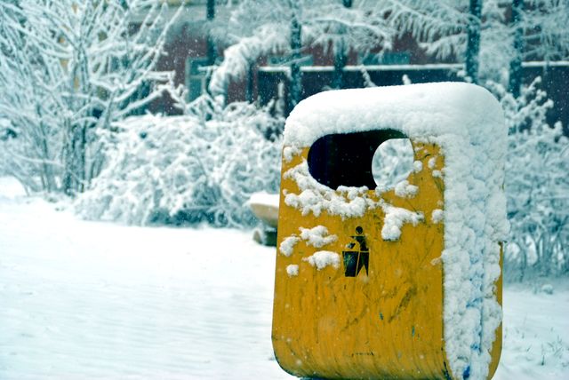 Yellow outdoor trash bin covered in snow, indicating a cold and frosty winter scene. Snow blankets the surroundings including trees and ground, setting a serene yet chilly atmosphere. Ideal for use in winter-themed projects, environmental campaigns, waste management promotions, or any context highlighting cold weather conditions.