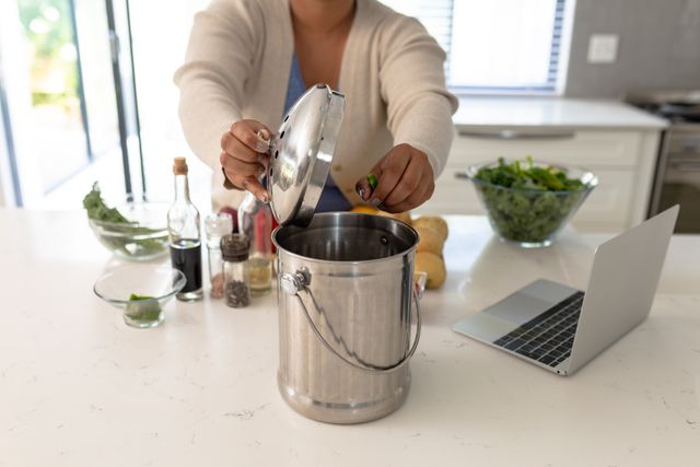 Midsection of an African American woman disposing of vegetable waste into a kitchen garbage bin. The scene includes various kitchen items like a laptop, spices, and fresh vegetables. This image is ideal for illustrating concepts related to sustainability, eco-friendly practices, waste management, and domestic life. It can be used in articles, blogs, and advertisements promoting green living, home organization, and cooking tips.