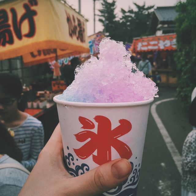 Person holding cup of colorful shaved ice with vibrant hues of blue and pink in focus. Background features blurred stalls and people enjoying a traditional Japanese festival. Perfect for use in articles about Japanese culture, travel blogs, food festivals, and vibrant street food experiences.