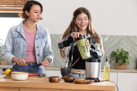Young women blending green smoothie while chopping tomatoes on wooden kitchen island