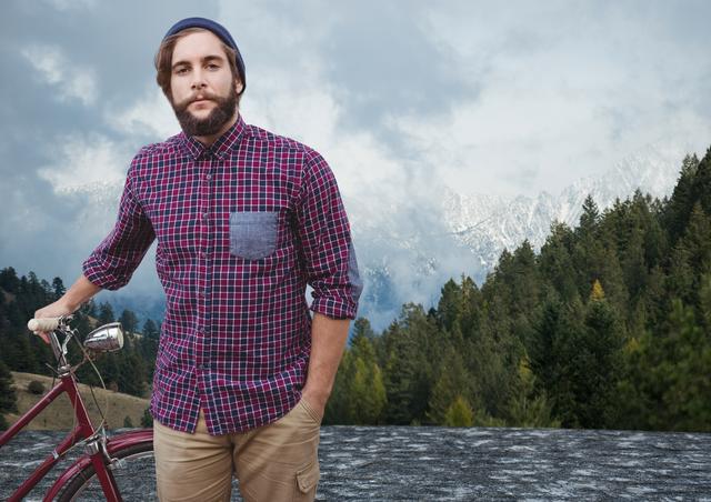This image depicts a man standing beside his bicycle with hands in pockets, wearing a plaid shirt and a beanie. The man is set against a picturesque background of mountains and lush coniferous forest. It can be used for content related to outdoor adventures, travel, nature, lifestyle blogging, and promoting casual and relaxed vibes.