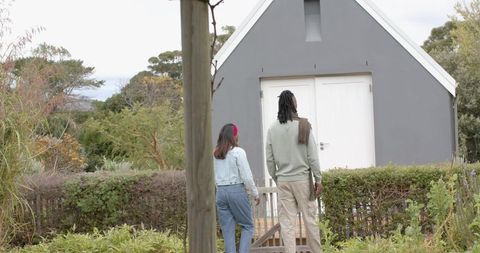Diverse couple walking toward modern gray garden outbuilding on short wooden deck near trimmed hedge