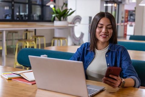 Young professional using laptop and smartphone in modern coworking lounge, smiling, focused